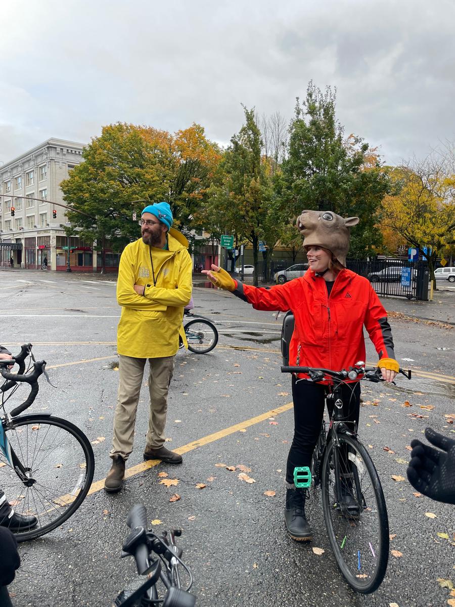 two people standing around talking, one sitting on a bike with a squirrel mask on